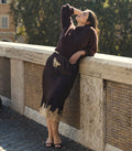 A woman leans against a stone bridge railing outdoors, wearing a dark brown knit sweater and matching midi skirt with beige floral embroidery and a beige lace hem.