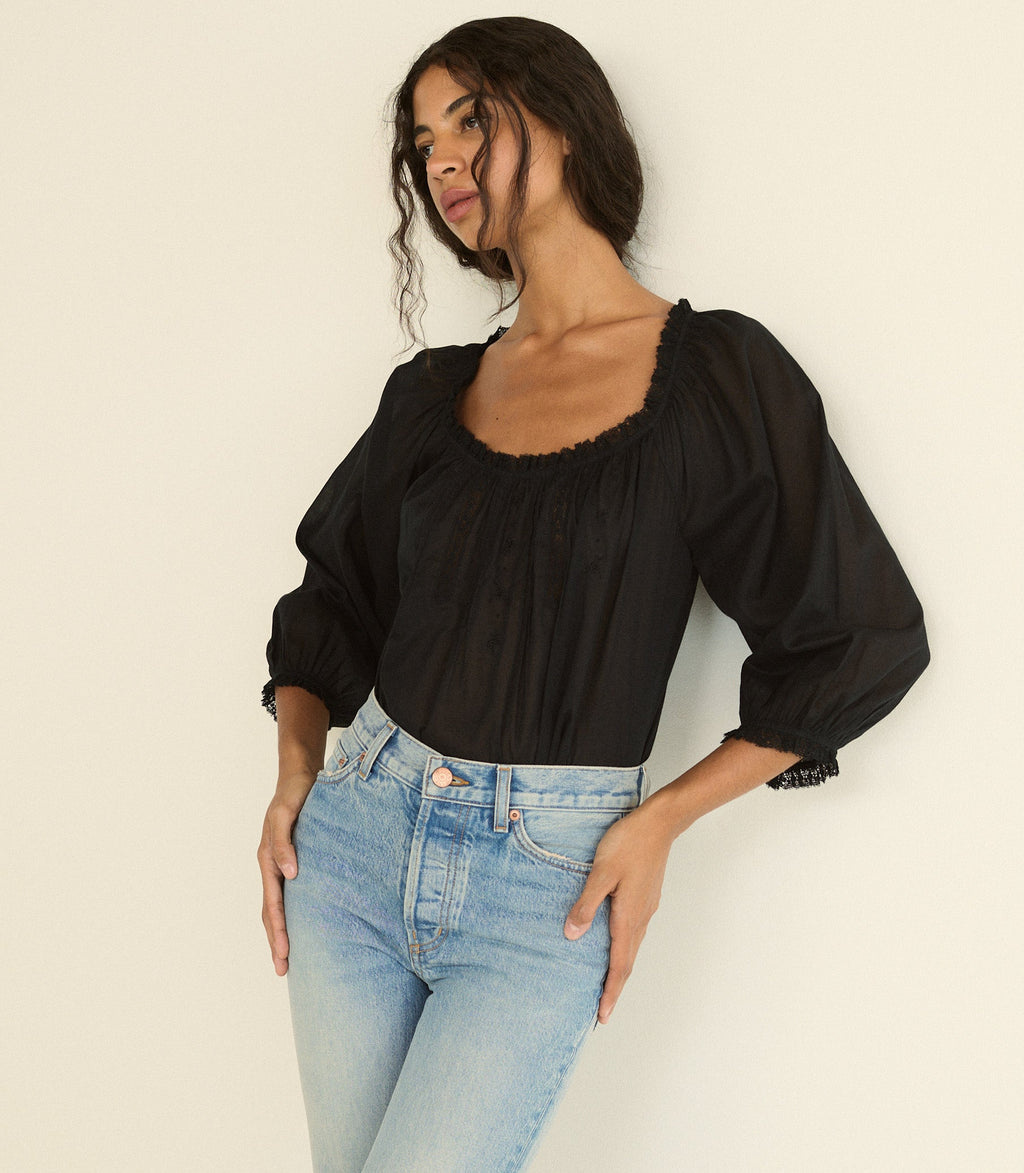 A woman wearing a black puff-sleeve blouse with a square neckline and scalloped trim, paired with light blue jeans, posing against a plain off-white background.