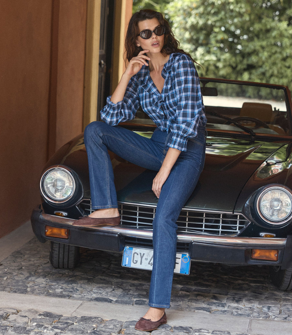 A woman wearing a blue and black plaid button-down shirt, dark blue jeans, and brown flat shoes sits on the front bumper of a vintage black car in an outdoor setting.