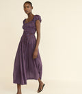 A woman modeling a midi-length purple dress with a small red and white foulard pattern, puffed cap sleeves, and a smocked bodice, set against a solid cream background.
