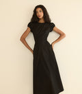 A woman modeling a black maxi dress with short puff sleeves, a round neckline, and a gathered waist, posing against a plain off-white background.