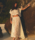 Woman in a white lace top and matching skirt standing against a rock with a natural background